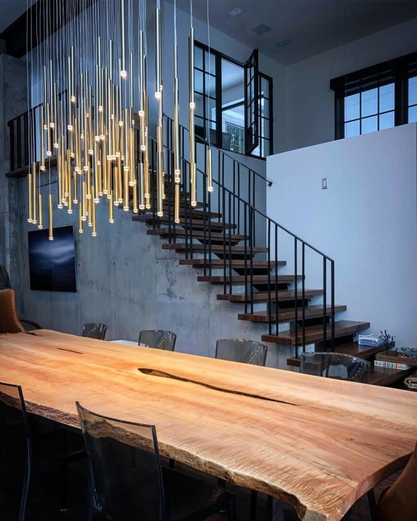 A modern dining room features a long wooden table with a natural edge, surrounded by transparent chairs. Above, numerous slender pendant lights hang in an artistic cluster. A staircase with black railings ascends in the background.