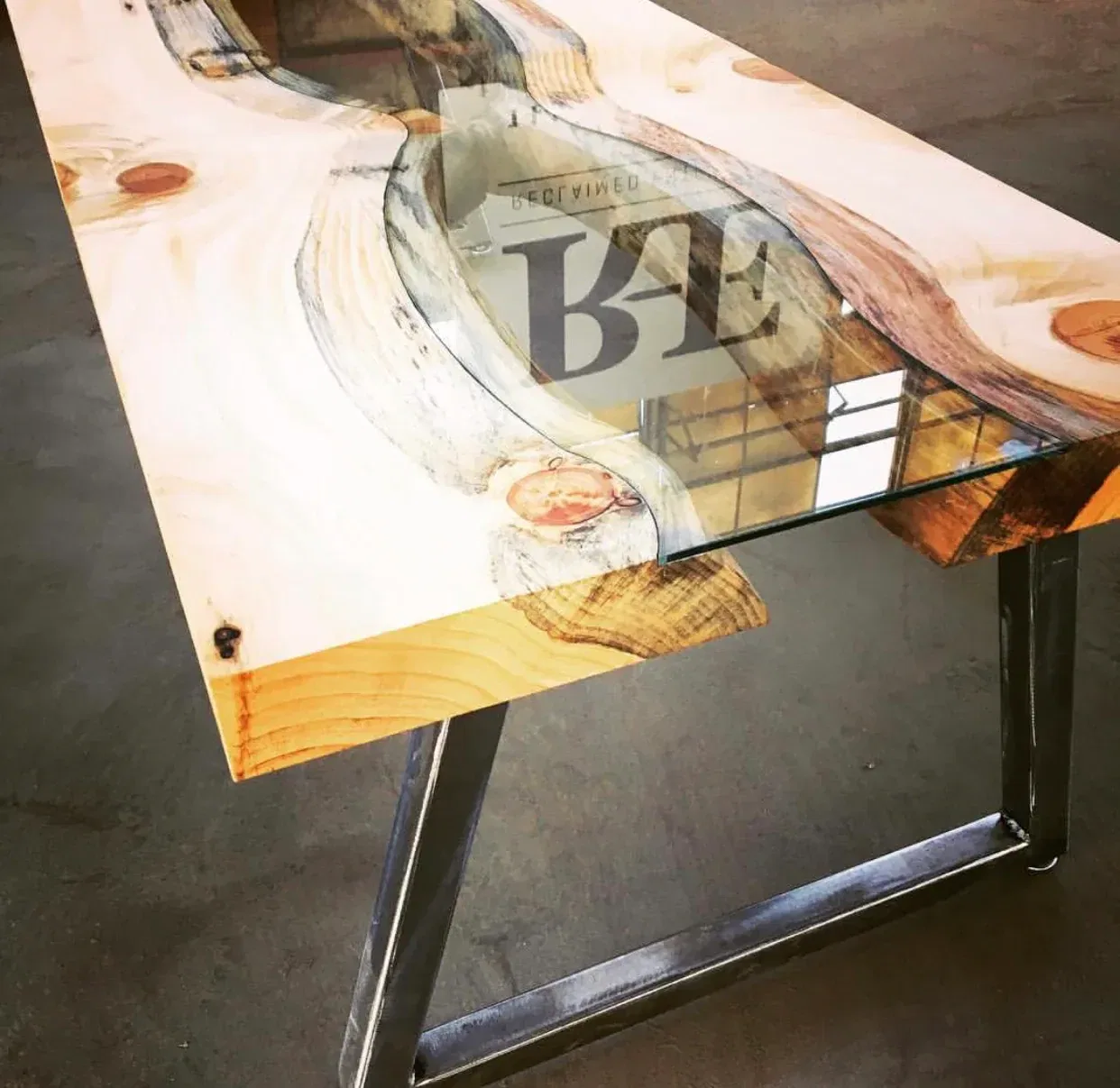 A wooden table with an intricate design featuring a river-like glass insert flows through the center. The table has metal legs, combining rustic and modern styles. The light-colored wood has distinct grain patterns and knots.