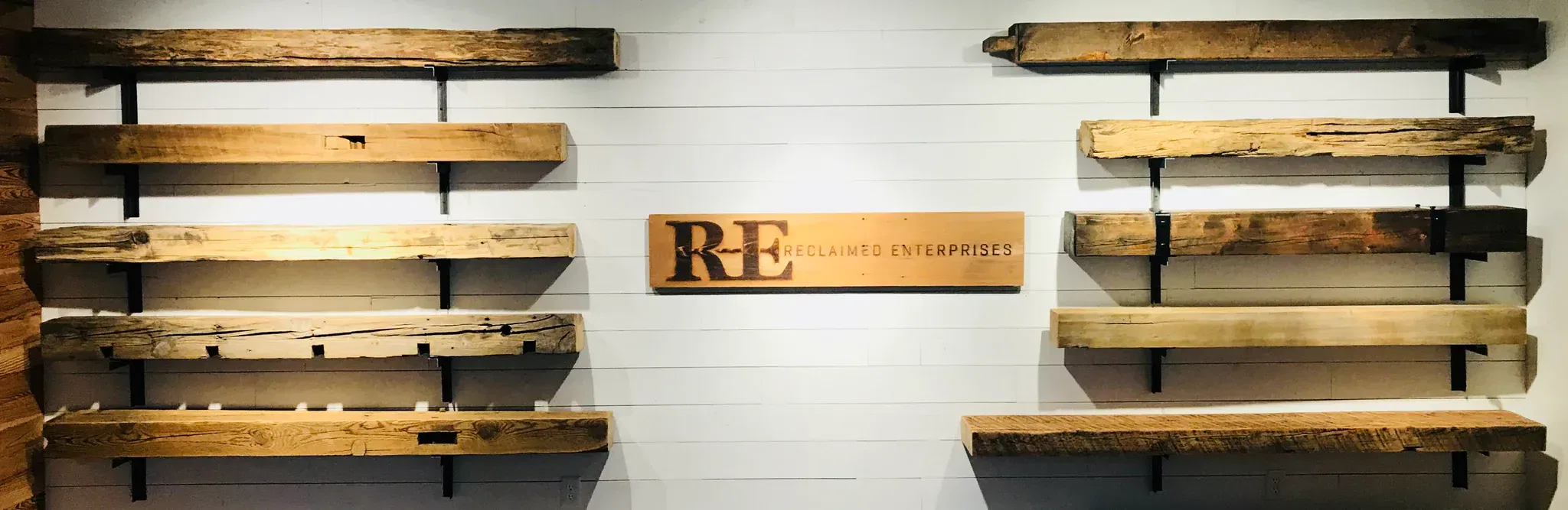 A display of rustic wooden shelves and mantels crafted from reclaimed wood, mounted on a white wall, features a central sign reading RE Reclaimed Enterprises. The Reclaimed Wood Mantels add a rugged, natural aesthetic to the setting.