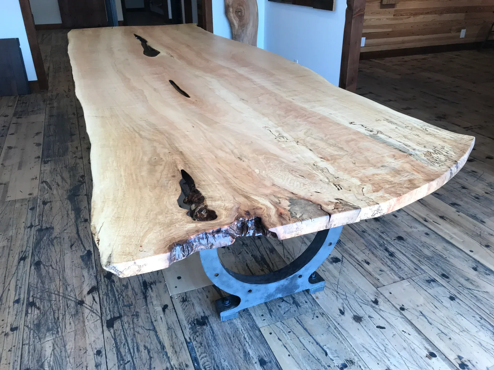 A large Live Edge Tops wooden table with a natural finish stands on a uniquely shaped metal base, featuring natural grain patterns and a few dark crevices. The floor and walls are crafted from rustic wood planks.