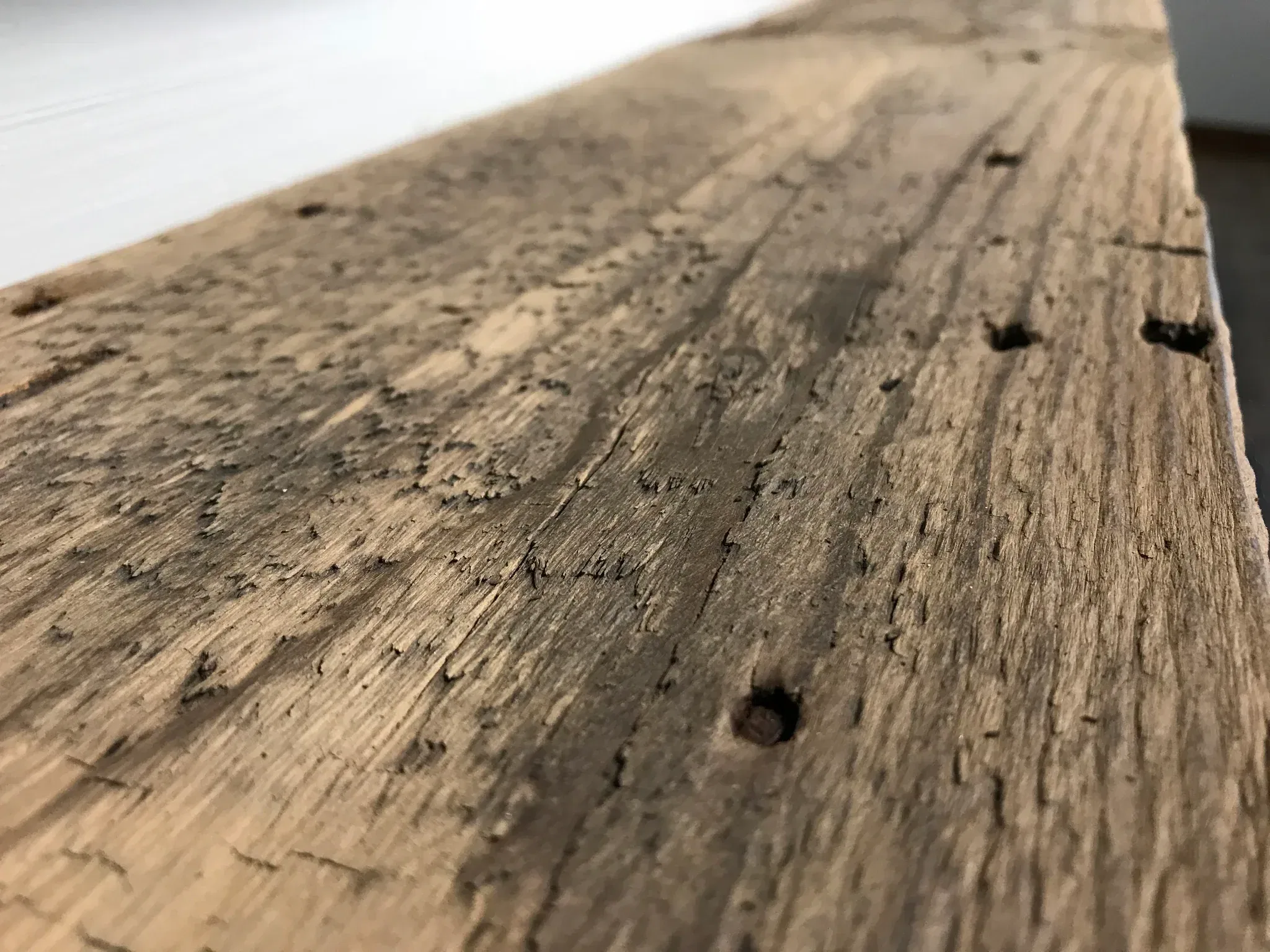 Close-up of a piece from the Reclaimed Wood Mantels collection, showcasing its weathered wooden surface with visible grain, small holes, and a rough texture that evokes the charm of antique mantels. The reclaimed wood has a rustic, aged appearance and stretches diagonally across the image against a blurred backdrop.