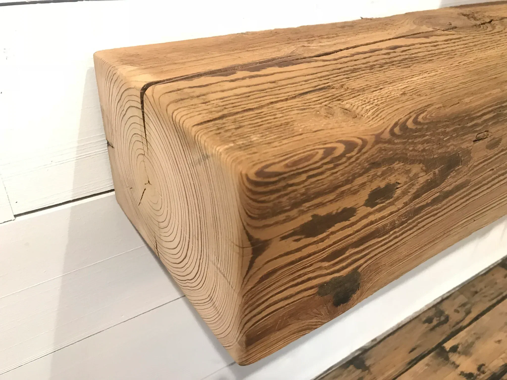 Close-up of a Reclaimed Wood Mantel mounted horizontally on a white wall, its rustic charm highlighted by visible grain, natural weathered texture, and knotholes. The floor beneath enhances the scene with its wooden finish.