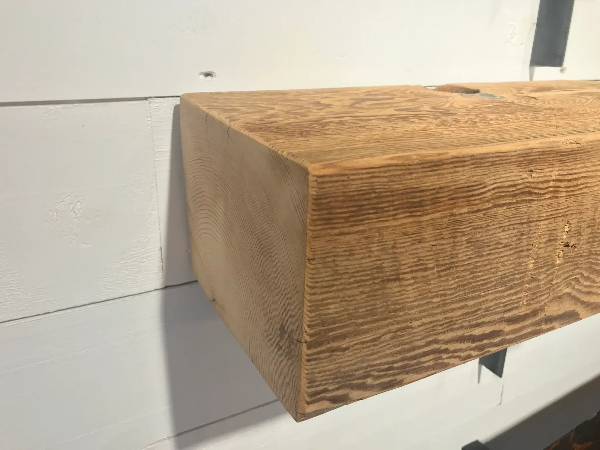 A close-up of a Reclaimed Wood Mantels beam extends from a white wall. Featuring natural grain texture and square edges, it is positioned horizontally and secured by metal brackets. Sunlight casts a subtle shadow on the wall.