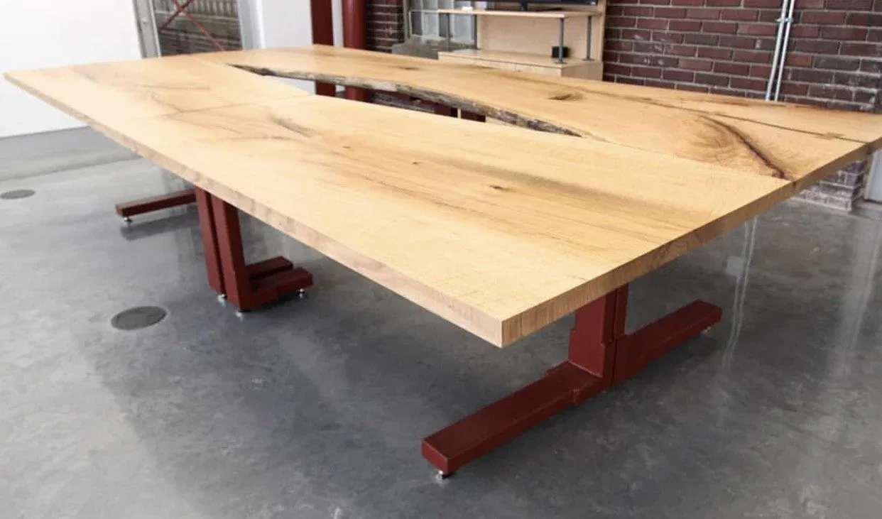 A large wooden table with a natural finish sits on red metal legs in a modern room with a concrete floor and brick walls. The tabletop features unique grain patterns and a central split design.