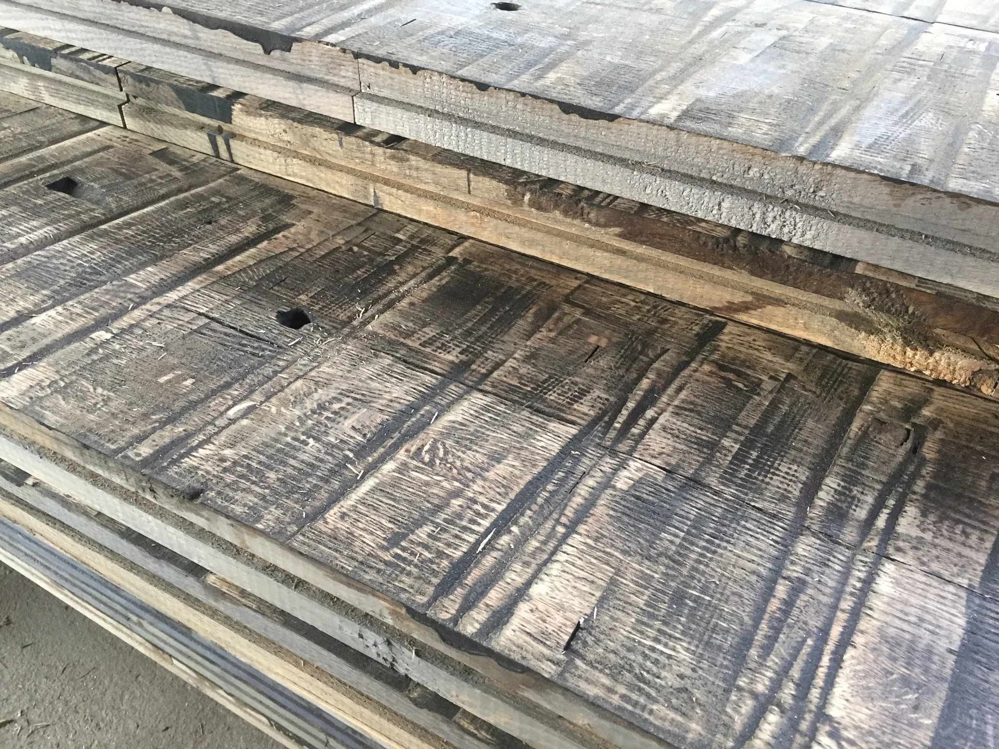A stack of Reclaimed Rail Car Plank boards with visible grain and cut marks lies on a surface, showcasing a weathered appearance with natural textures and shades of brown and gray.