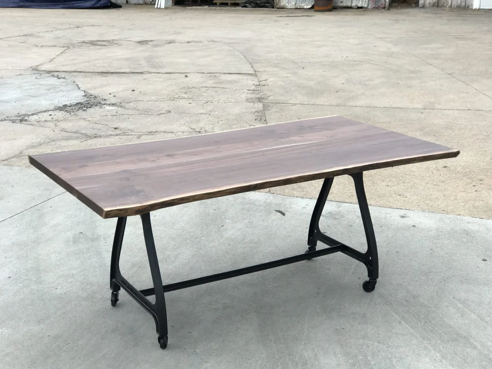 A wooden table with a smooth, rectangular top and natural edges stands on a metal frame with four wheels, placed on a concrete surface. The background is an open outdoor area.