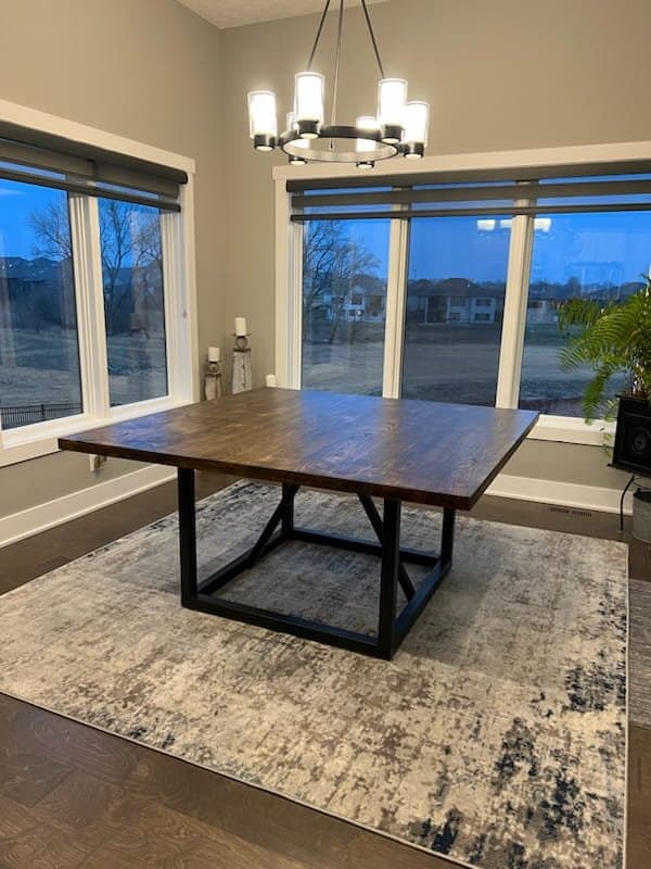 A dining room with a square wooden table on a patterned rug. Large windows provide a view of the outside, and a modern chandelier hangs above the table. The room features neutral colors and a potted plant in the corner.
