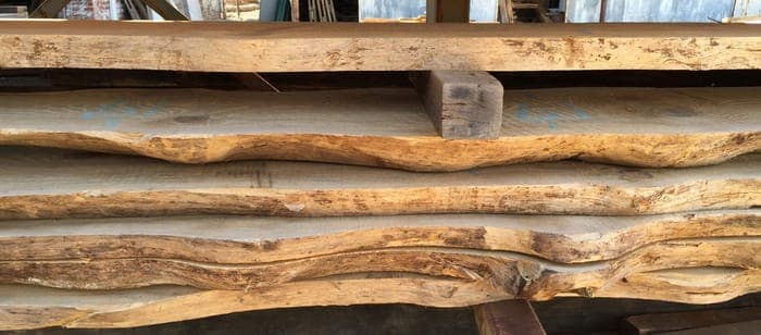 Stacked slabs of raw, unfinished wood with irregular, natural edges are neatly arranged. The wood appears to be in a workshop setting, hinting at carpentry or woodworking projects.