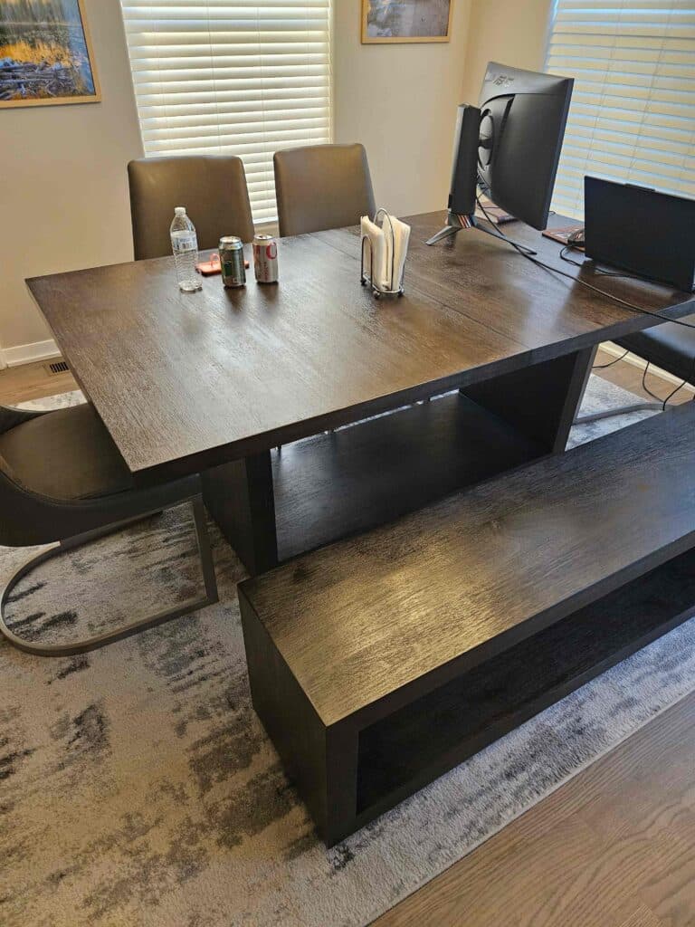 A dark wooden dining table with a matching bench and chairs, set with drinks, a water bottle, napkins, and a computer monitor, in a room with light-colored walls and windows with blinds.