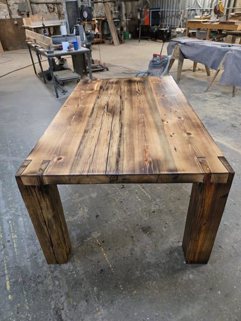 A handcrafted wooden table with a rustic finish stands in a workshop, surrounded by tools, workbenches, and equipment. The table features thick legs and a polished, textured tabletop with visible wood grain and knots.