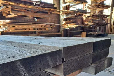 Stacks of large, weathered wooden beams are neatly piled in a warehouse. Sunlight streams in from windows, highlighting the rough and textured surfaces. The beams are arranged on a pallet, emphasizing the rustic, industrial setting.