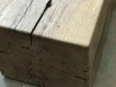 Close-up of a textured wooden beam with visible cracks and natural wood grain. The beam is situated on a light surface near a window, providing soft natural lighting.