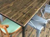 A wooden table with metal legs is surrounded by various chairs, including an upholstered wooden chair with a green cushion and several gray metal chairs. The floor has a rustic, scratched wooden appearance.