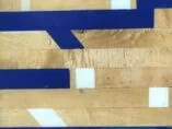 Close-up of a gymnasium floor showcasing various shades of wooden parquet with blue and white painted lines forming geometric patterns across the surface. The lines create a contrast against the wood grain.
