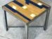 A small square table with a wooden top featuring a geometric design in blue and natural wood tones. The table has a metal frame and legs, and it is placed on a gray concrete floor.