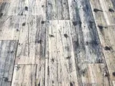 A close-up view of a distressed wooden floor with a pattern of dark stains, scratches, and wear, highlighting the rustic and weathered texture of the planks.