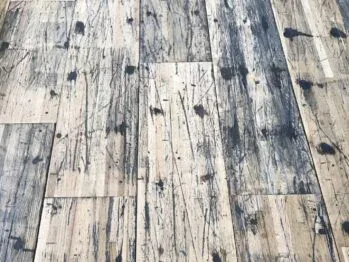 A close-up view of a distressed wooden floor with a pattern of dark stains, scratches, and wear, highlighting the rustic and weathered texture of the planks.