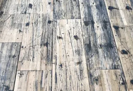 A close-up view of a distressed wooden floor with a pattern of dark stains, scratches, and wear, highlighting the rustic and weathered texture of the planks.