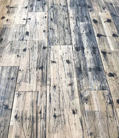 A close-up view of a distressed wooden floor with a pattern of dark stains, scratches, and wear, highlighting the rustic and weathered texture of the planks.