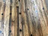 A close-up of a wooden table with a rustic appearance. The surface has a weathered texture with visible wood grain and scattered dark circular markings. The color varies from light to dark brown tones.