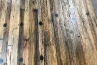 A close-up of a wooden table with a rustic appearance. The surface has a weathered texture with visible wood grain and scattered dark circular markings. The color varies from light to dark brown tones.