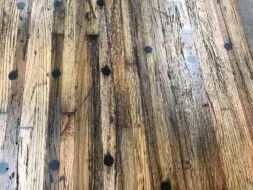 A close-up of a wooden table surface featuring a rustic design with visible grain patterns and scattered black circular markings. The wood appears weathered and textured, contributing to its vintage look.