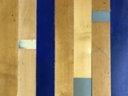 Close-up of a gymnasium floor, featuring alternating wooden planks in light brown and blue, with gray and beige patches on some planks. The surface shows signs of wear and visible seams.