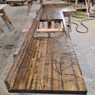A long, L-shaped wooden bench under construction in a workshop. The surface appears worn and marked with visible scratches and cuts. In the background, there are tools, lumber, and coiled cables.