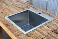 A square metal sink is installed in a rustic wooden countertop. The background features a corrugated metal wall, adding an industrial feel to the setting.