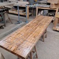 A long wooden table with a rustic, textured surface is set in a woodworking shop. Various tools and pieces of wood are in the background, placed on shelves and workbenches. The space has an industrial, handcrafted feel.