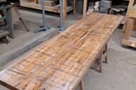 A long wooden table with a rustic, textured surface is set in a woodworking shop. Various tools and pieces of wood are in the background, placed on shelves and workbenches. The space has an industrial, handcrafted feel.