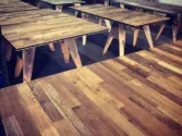 A workshop filled with several handcrafted wooden tables, each featuring a unique pattern of mixed wood tones. The tables are arranged in rows, highlighting their rustic, artisanal design. The background shows tools and workbenches.