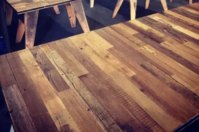 A workshop filled with several handcrafted wooden tables, each featuring a unique pattern of mixed wood tones. The tables are arranged in rows, highlighting their rustic, artisanal design. The background shows tools and workbenches.