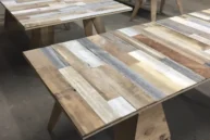 Wooden tables with plank-style surfaces and sawhorse legs are arranged in a workshop. Each table features a variety of wood tones and textures, creating a rustic and handcrafted appearance.
