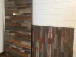 Two weathered wooden panels with a mix of brown, gray, and blue tones lean against a white shiplap wall on a wooden floor.