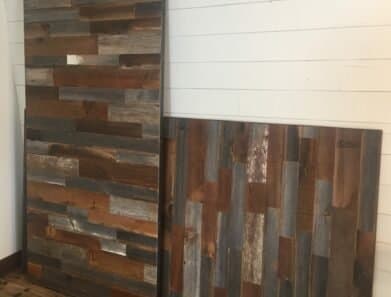 Two weathered wooden panels with a mix of brown, gray, and blue tones lean against a white shiplap wall on a wooden floor.