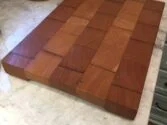 A wooden cutting board with a checkered pattern of alternating light and dark wood grains sits on a marble countertop. The board features square blocks, emphasizing the natural wood textures.