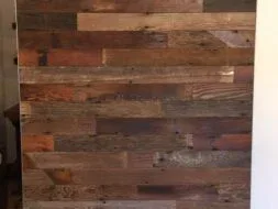 A wooden sliding barn door with a mix of dark and light brown planks, mounted on a black metal rail system, set against a white wall. The wood grain and texture are visible, giving a rustic appearance.