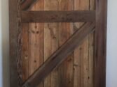 A rustic wooden barn door with a diagonal Z-brace design, made of weathered, vertically aligned wooden planks. It hangs on a black metal sliding track above a wooden floor. The door exudes a vintage, farmhouse aesthetic.