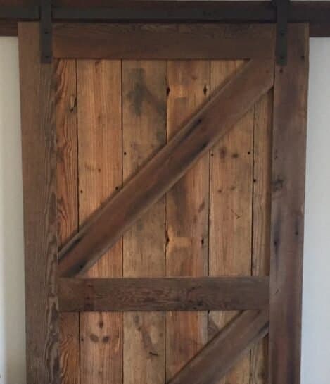 A rustic wooden barn door with a diagonal Z-brace design, made of weathered, vertically aligned wooden planks. It hangs on a black metal sliding track above a wooden floor. The door exudes a vintage, farmhouse aesthetic.