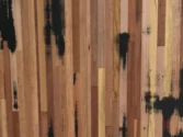 Wooden flooring with vertical planks of varying shades of brown, featuring black stains scattered across the surface. The texture and natural grain of the wood are visible, creating a rustic and artistic pattern.