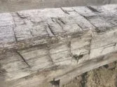 Close-up of a weathered wooden beam with visible cracks and a small hole in the center. The surface appears rough and aged, resting on a sandy ground.
