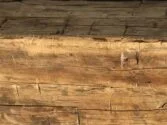 Close-up of a weathered wooden beam with a rough, textured surface. The wood displays signs of aging, including cracks and variations in color, creating an antique appearance.
