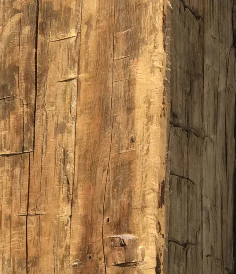 Close-up of a wooden beam showing natural textures, grooves, and some weathering. The woods warm brown tone and visible grain patterns suggest an aged, rustic appearance.
