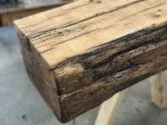 Close-up of a rustic wooden beam with visible grain and natural imperfections. The beam has a textured, weathered appearance, highlighting the rough edges and rich wood tones. It rests on a wooden surface.