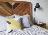 A cozy bedroom with a wooden headboard, geometric-patterned bedding in gray and yellow tones, and a matching pillow. A black wall-mounted lamp is above a wooden nightstand, which holds a white mug and a small notepad with a pen.