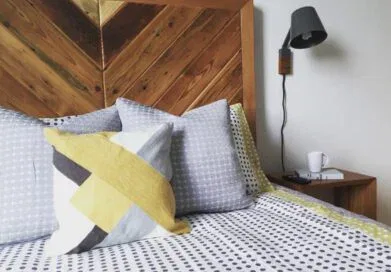 A cozy bedroom with a wooden headboard, geometric-patterned bedding in gray and yellow tones, and a matching pillow. A black wall-mounted lamp is above a wooden nightstand, which holds a white mug and a small notepad with a pen.