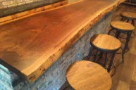 A Live Edge Tops wooden bar counter with a natural finish is set against a brick wall. Four round wooden stools with metal frames are neatly aligned along the bar, which sits atop a floor made of wood planks.