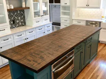 A modern kitchen with a long wooden island countertop. The island has teal cabinets and stainless steel appliances. Surrounding white cabinetry features open shelves, a black and white decorative backsplash, and a tile backsplash near the sink.