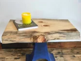 A small wooden table with a blue metal base sits on a wooden floor. A yellow candle and a book titled prattle rest on the tables surface, which features natural wood knots and grain patterns.